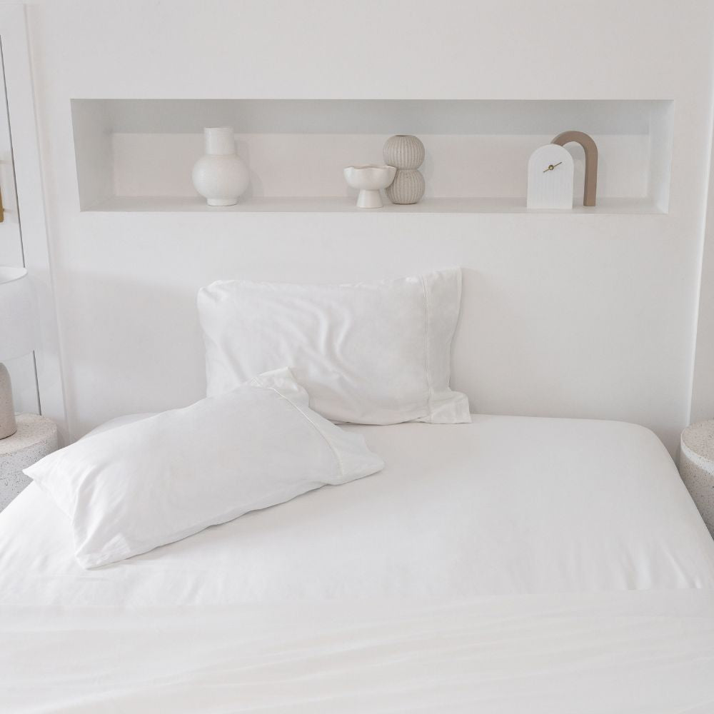 A neatly made bed with white sheets and two pillows in a minimalistic white room. A recessed shelf above the bed holds decorative white vases and small sculptures.