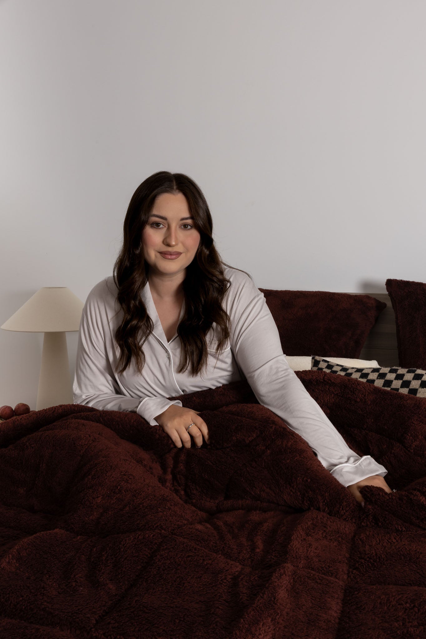 Woman sitting on a couch under a dark brown blanket in a room with a lamp and checkered pillow.