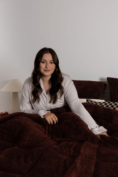 Woman sitting on a couch under a dark brown blanket in a room with a lamp and checkered pillow.