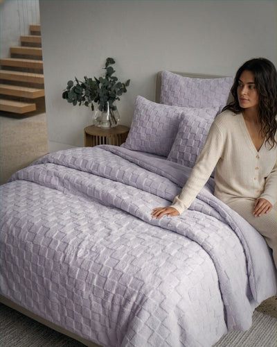 Lifestyle shot of lavender checkered fleece comforter set on bed with woman in cream cardigan seated beside it.