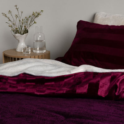 Red bedding with a white blanket on a bed, next to a wooden side table with a vase and water pitcher.