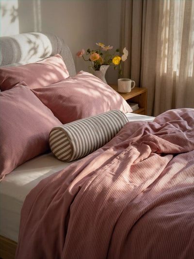 Pink bedding with striped pillow on a bed in a softly lit room.