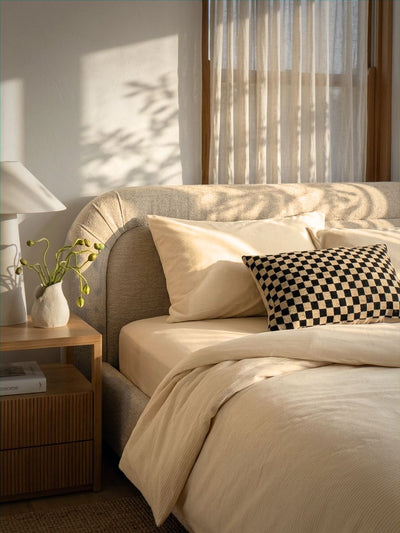 A neatly made bed with cream bedding and a black-and-white checkered accent pillow, shown in a softly lit bedroom with sunlight, sheer curtains, a bedside table, and a lamp.