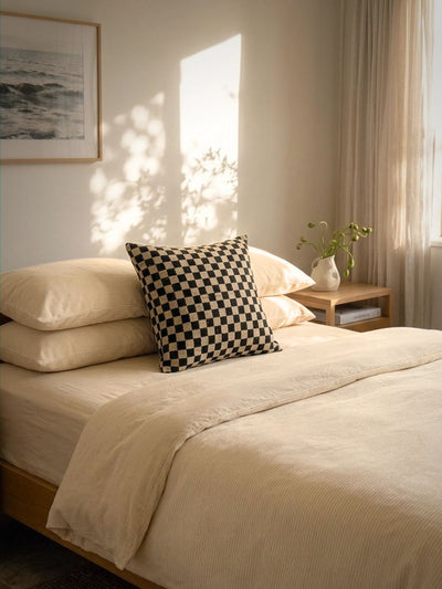A neatly made bed with cream bedding and a black-and-white checkered accent pillow in a softly lit bedroom. A wooden nightstand with a small vase of greenery sits beside the bed, and warm sunlight streams across the wall.