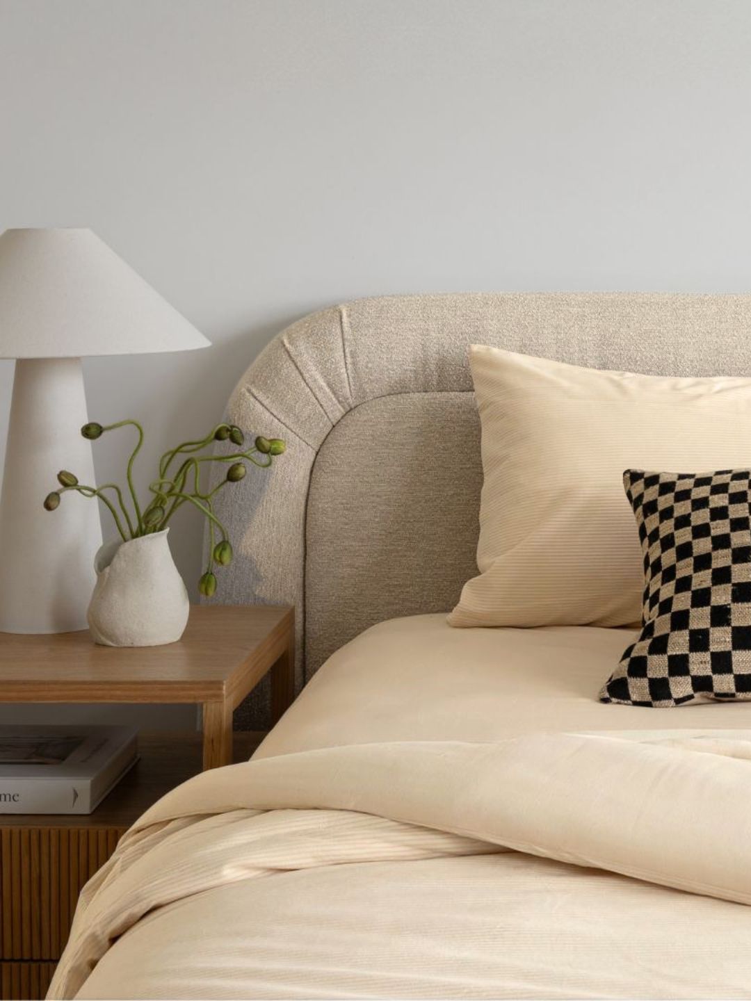 A close-up of a neatly made bed with cream bedding, a beige upholstered headboard, a white bedside table, a white lamp, and a small black-and-white checkered accent pillow against a light gray wall.