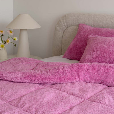 Pink textured blanket and pillows on a bed with a lamp and flowers in the background.