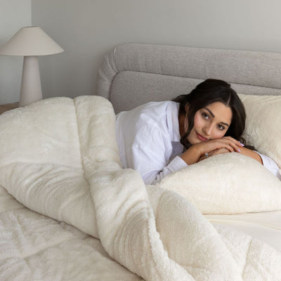 Woman in a robe lying in bed under a white comforter, with a lamp in the background.