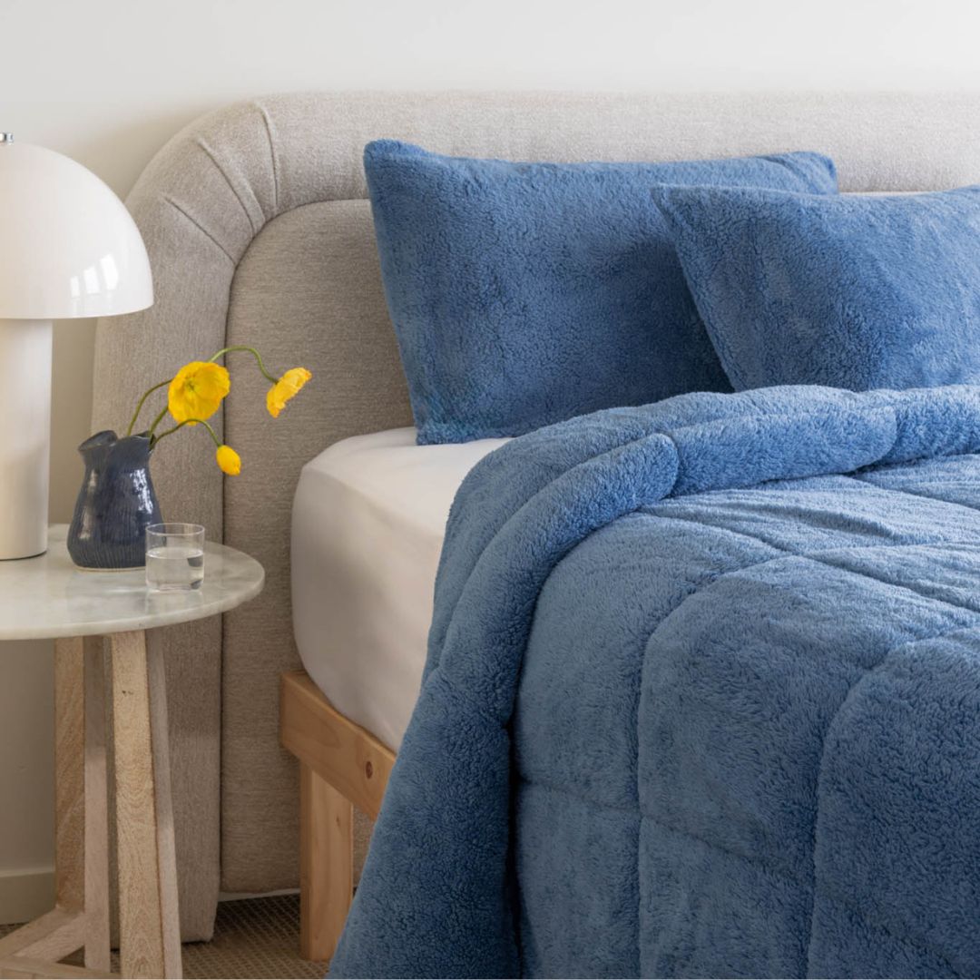 Blue textured blanket and pillows on a bed with a side table and lamp in a bedroom setting.