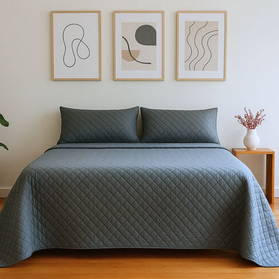 A neatly made bed features the Morgan and Reid Argyle Shade Coverlet Set in dark gray with matching pillows, creating a contemporary vibe. Above, three abstract art pieces hang, while a wooden table with red flowers sits to the right.