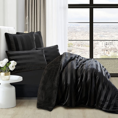 A modern bedroom features the Morgan and Reid Black Velvet Stripe Snuggle Fleece Comforter Set on a neatly made bed with striped pillows, neutral walls, abstract art, a beige chair, round black side table with white flowers, and a lamp.