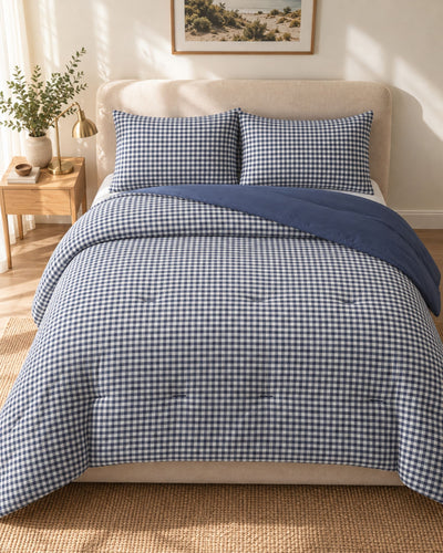 A neatly made bed in a sunlit bedroom featuring a blue-and-white gingham check comforter, matching pillow shams, and a solid navy throw blanket draped across the top. A beige upholstered headboard, wood nightstand, plant, and framed wall art are visible in the room.