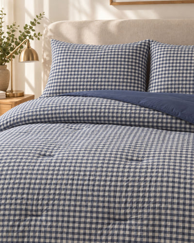 A neatly made bed with a blue and white gingham check comforter, matching pillow shams, and a solid navy blue folded back section, shown in a softly lit bedroom setting with a headboard, bedside lamp, and plant.
