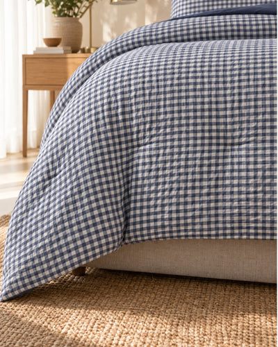 A close-up of a bed dressed in a blue and white gingham checkered comforter, with matching pillows, a beige upholstered bed frame, a woven jute rug, and a wooden side table in a softly lit bedroom.
