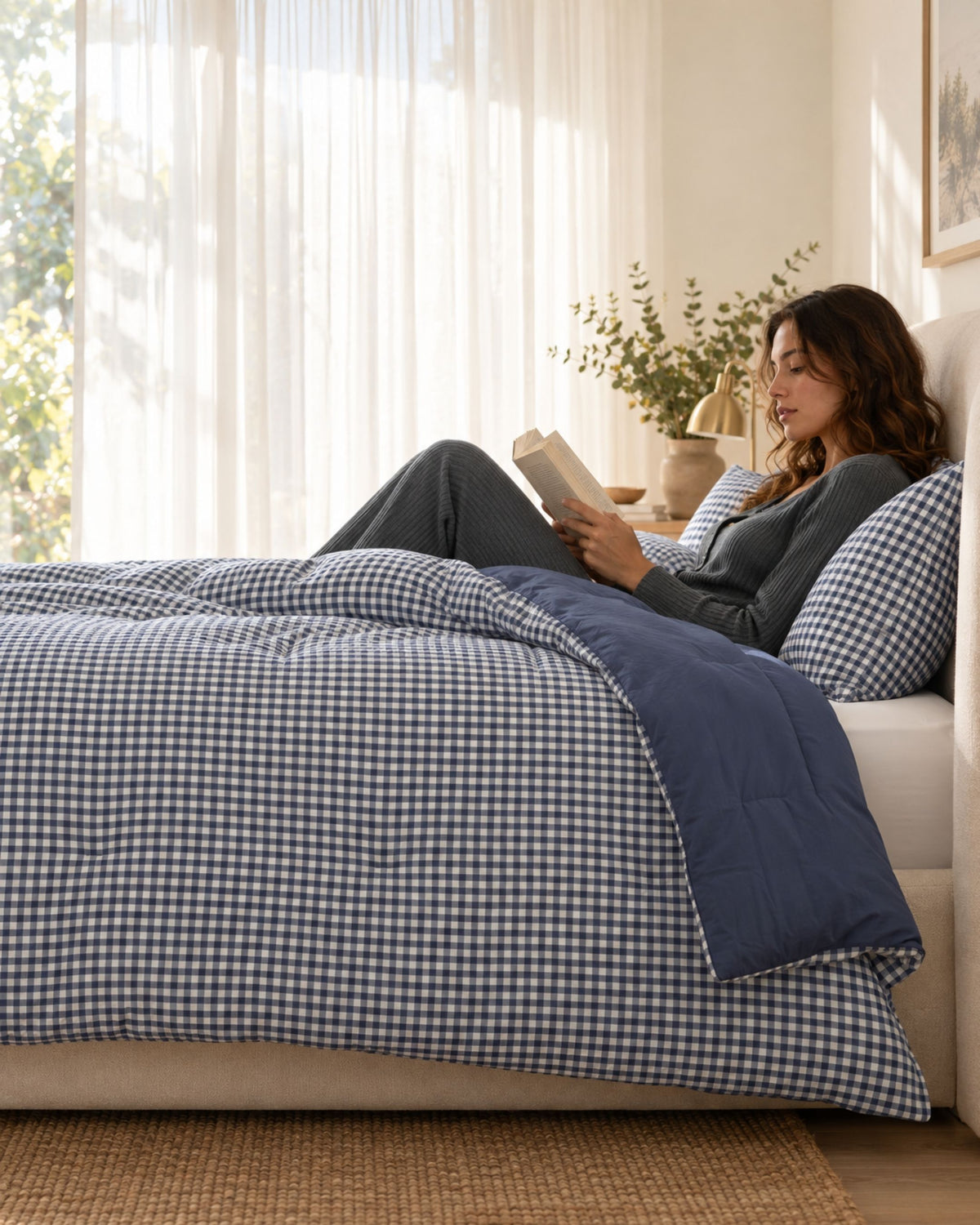 A woman relaxes on a bed in a bright bedroom, reading a book. The bed is made with a navy and white gingham checkered duvet cover and matching pillowcases, with a solid navy blue quilted throw blanket.