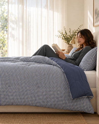 A woman relaxes on a bed in a bright bedroom, reading a book. The bed is made with a navy and white gingham checkered duvet cover and matching pillowcases, with a solid navy blue quilted throw blanket.