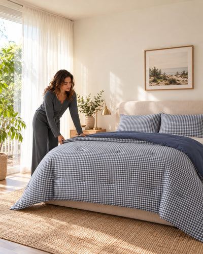 A woman in a gray long-sleeve dress is adjusting a blue and white gingham checkered comforter on a bed in a bright, sunlit bedroom. The bed also has matching pillow shams, a beige upholstered headboard, a woven jute rug, and a framed landscape print on the wall.