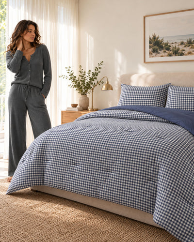 A woman stands beside a bed in a softly lit bedroom wearing a matching gray cardigan and wide-leg pajama pants. The bed is made with a blue-and-white gingham comforter set, matching pillows, and a solid navy throw blanket.