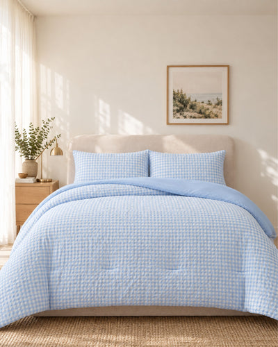 A neatly made bed in a bright bedroom, covered with a light blue and white gingham quilt and matching pillow shams. The room has warm cream walls, a beige upholstered headboard, a small wooden nightstand with a lamp and vase, and a framed landscape print above the bed.