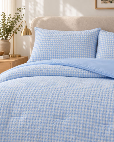 Light blue and white gingham patterned bedding set on a bed, including a comforter and two matching pillowcases, styled in a bright bedroom setting with a wooden nightstand and gold lamp.
