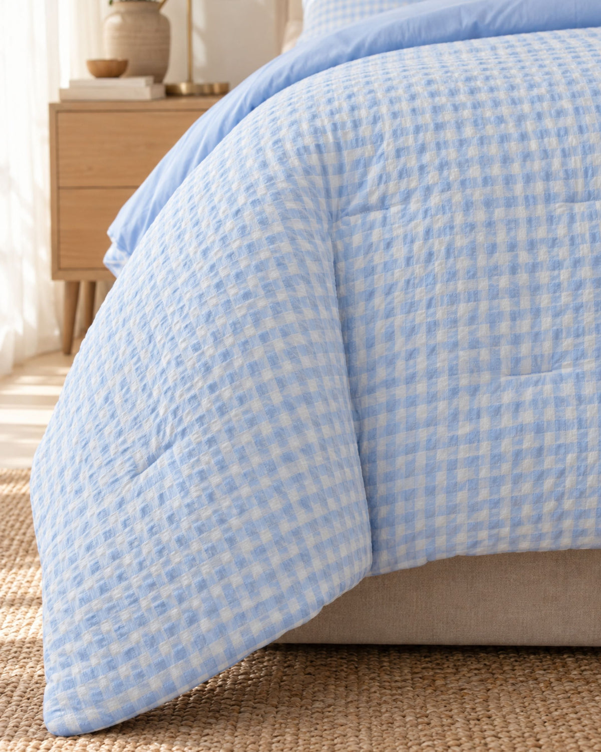 A light blue and white gingham patterned quilted comforter draped over a bed in a softly lit bedroom setting.