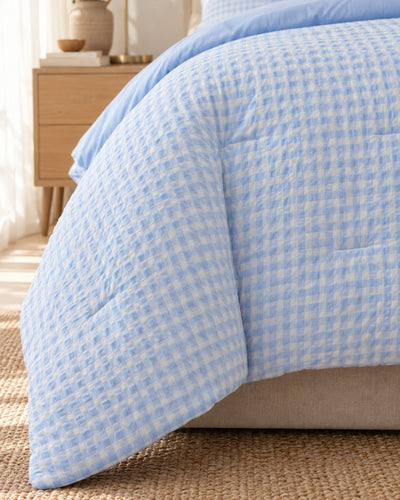 A light blue and white gingham patterned quilted comforter draped over a bed in a softly lit bedroom setting.