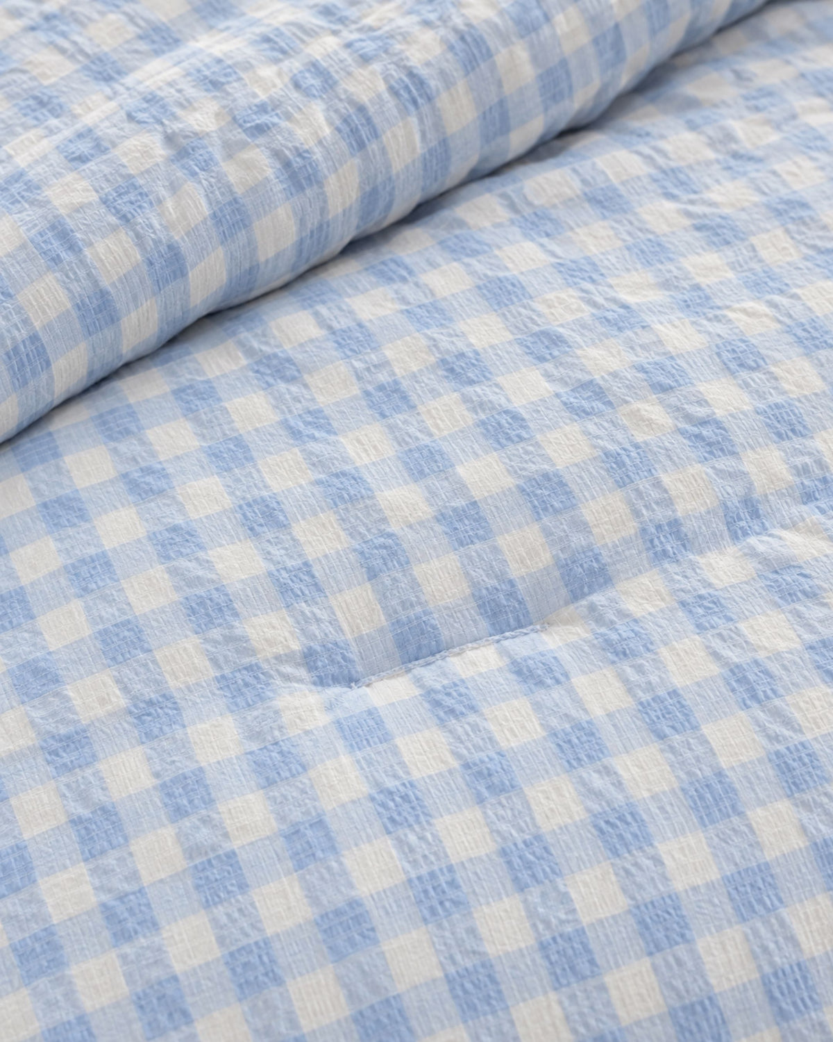 A close-up of a light blue and white gingham patterned fabric, likely a quilted comforter or duvet, showing its soft texture and folded corner.