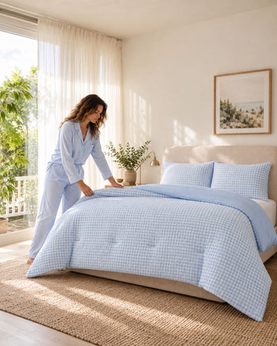 A woman in light blue pajamas adjusts a light blue and white gingham checkered comforter on a bed in a bright, sunlit bedroom.