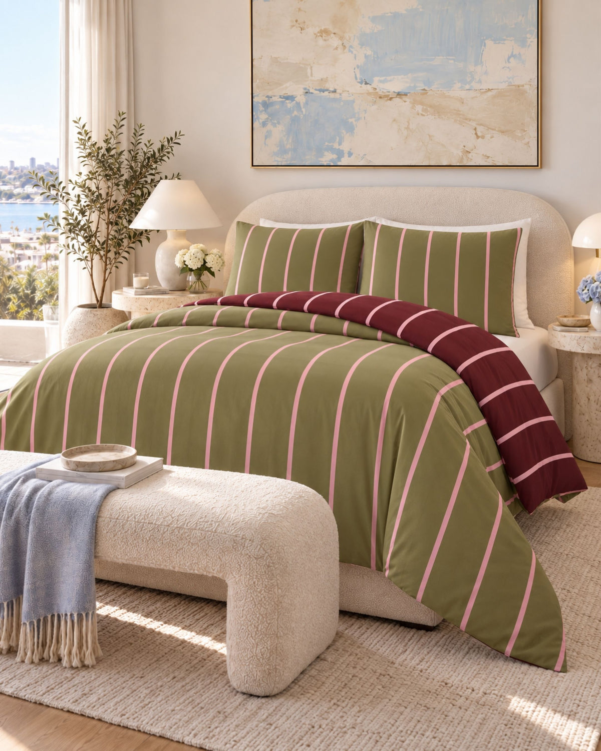 A styled bedroom scene showing a bed made up with an olive green duvet cover and matching pillow shams featuring vertical pink stripes, plus a folded burgundy striped throw. A cream bench, lamps, plant, and framed wall art are visible in the room.