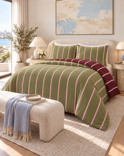 A styled bedroom scene showing a bed made up with an olive green striped bedding set, including a duvet cover and matching pillow shams, with a burgundy throw folded at the foot. The room includes a cream upholstered bench with a blue throw blanket, bedside tables and lamps, a window view, and wall art.