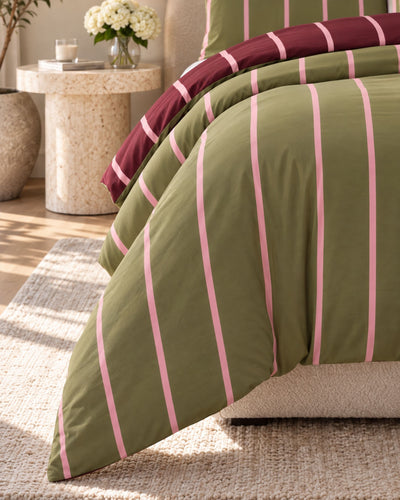 A close-up of a striped bedding set on a bed in a styled bedroom. The duvet cover is olive green with vertical pink stripes, and the folded top layer shows a dark burgundy side with pink stripes.