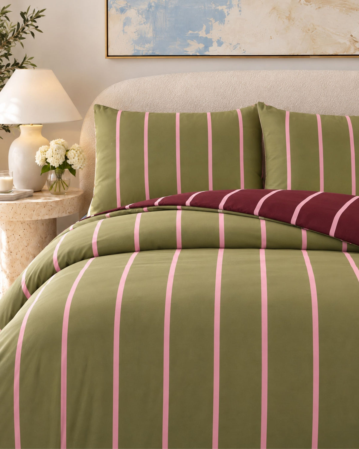 A bed dressed in olive green and pink vertically striped bedding, with two matching pillowcases and a folded burgundy striped throw blanket, shown in a styled bedroom setting with a bedside lamp and framed wall art.