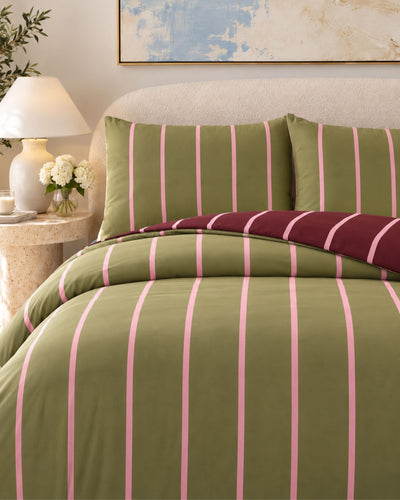 A bed dressed in olive green and pink vertically striped bedding, with two matching pillowcases and a folded burgundy striped throw blanket, shown in a styled bedroom setting with a bedside lamp and framed wall art.