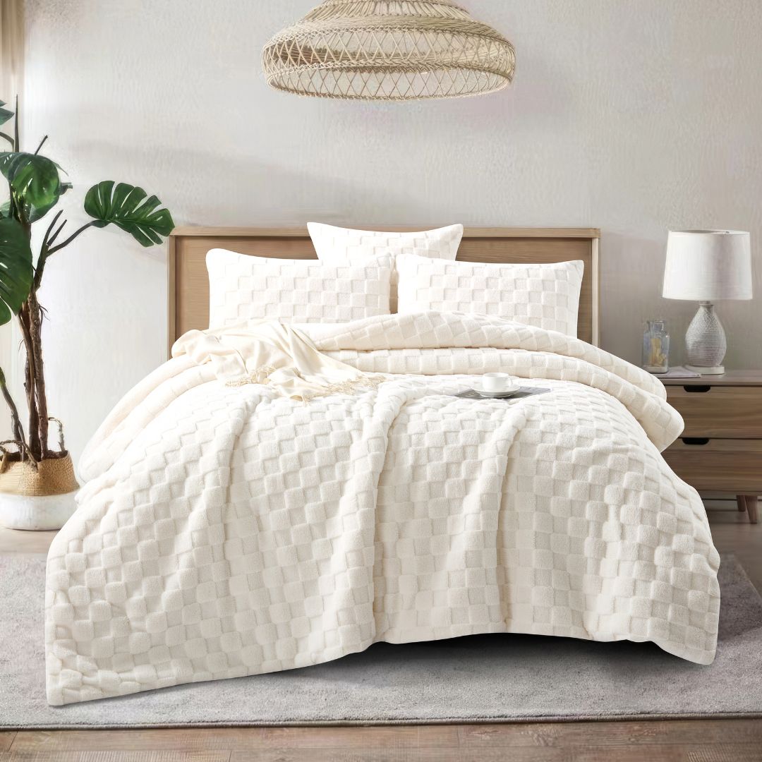 A neatly made bed showcases the Morgan and Reid Checkered Cream Snuggle Fleece Comforter Set with textured pillows, against a light wall, paired with a wooden headboard, bedside table, lamp, and a large leafy plant.