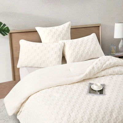 A neatly made bed showcases the Morgan and Reid Checkered Cream Snuggle Fleece Comforter Set with textured pillows, against a light wall, paired with a wooden headboard, bedside table, lamp, and a large leafy plant.