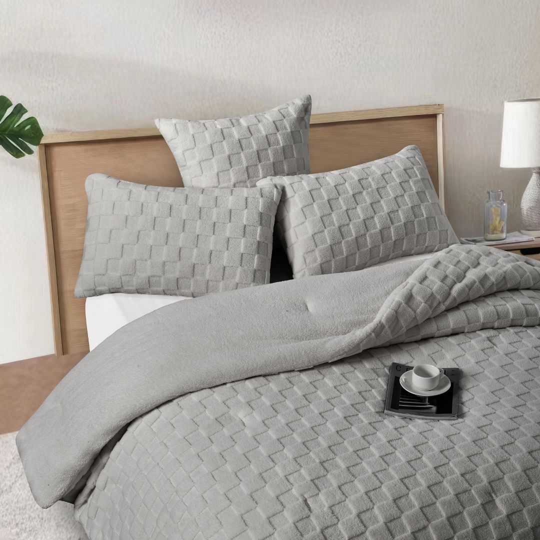 A neatly made bed features the Morgan and Reid Checkered Silver Snuggle Fleece Comforter Set, with matching pillows, a wooden headboard, nightstand with lamp and bottles, a large plant, and a woven pendant light for lasting luxury.