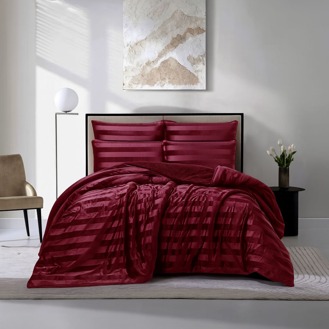 A modern bedroom features a Morgan and Reid Cherry Red Velvet Stripe Snuggle Fleece Comforter Set with burgundy pillows on a neatly made bed. Nearby are a beige chair, round lamp, white flowers, and abstract art on the light gray wall.