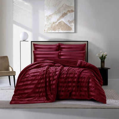 A modern bedroom features a Morgan and Reid Cherry Red Velvet Stripe Snuggle Fleece Comforter Set with burgundy pillows on a neatly made bed. Nearby are a beige chair, round lamp, white flowers, and abstract art on the light gray wall.