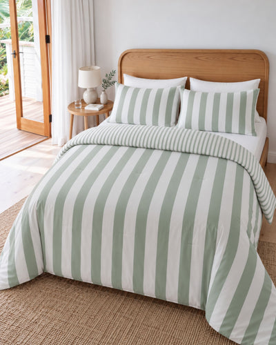 A bed dressed in a mint green and white striped duvet cover and matching pillow shams, styled in a bright bedroom with a wooden headboard, side table, lamp, and window looking out onto a deck.