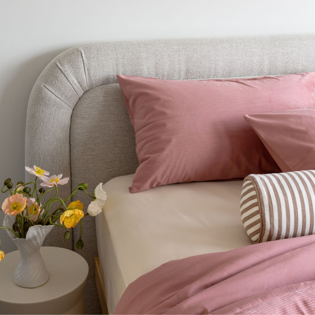 Bed with pink bedding and pillows, next to a small table with flowers, in a bedroom setting.