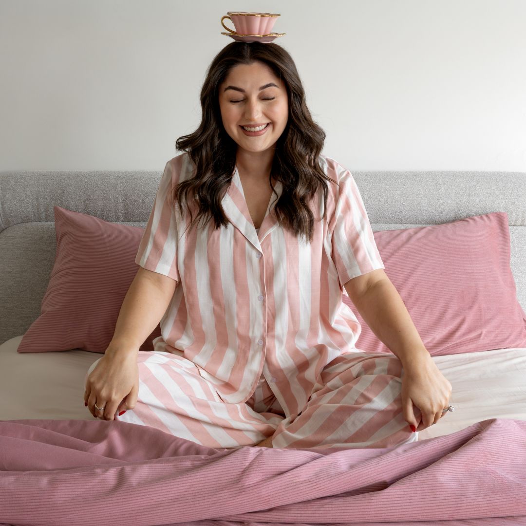 Woman in pink striped pajamas sitting on a bed with a teacup on her head.