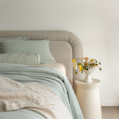 Neatly made bed with light bedding and pillows, next to a small table with a vase of flowers.