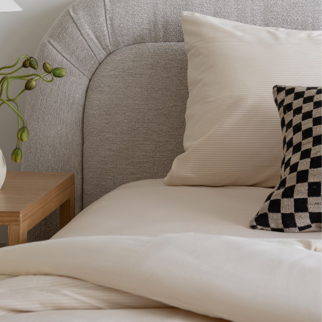 Close-up of a bed with beige pillows and a checkered pillow, wooden nightstand with plant, and gray upholstered headboard.
