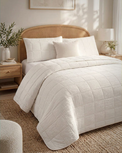 A neatly made bed in a softly lit bedroom, dressed with an off-white quilted comforter and matching shams. The bed has a natural woven rattan headboard, flanked by wooden nightstands with lamps and plants.