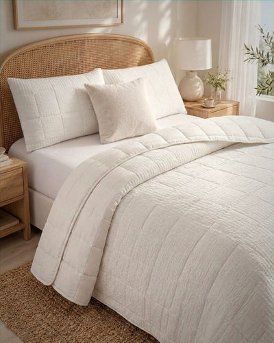 A neatly made bed in a bright bedroom setting, featuring a textured off-white quilted comforter, two matching pillow shams, a woven cane headboard, and light wood bedside tables with a lamp and small plant.