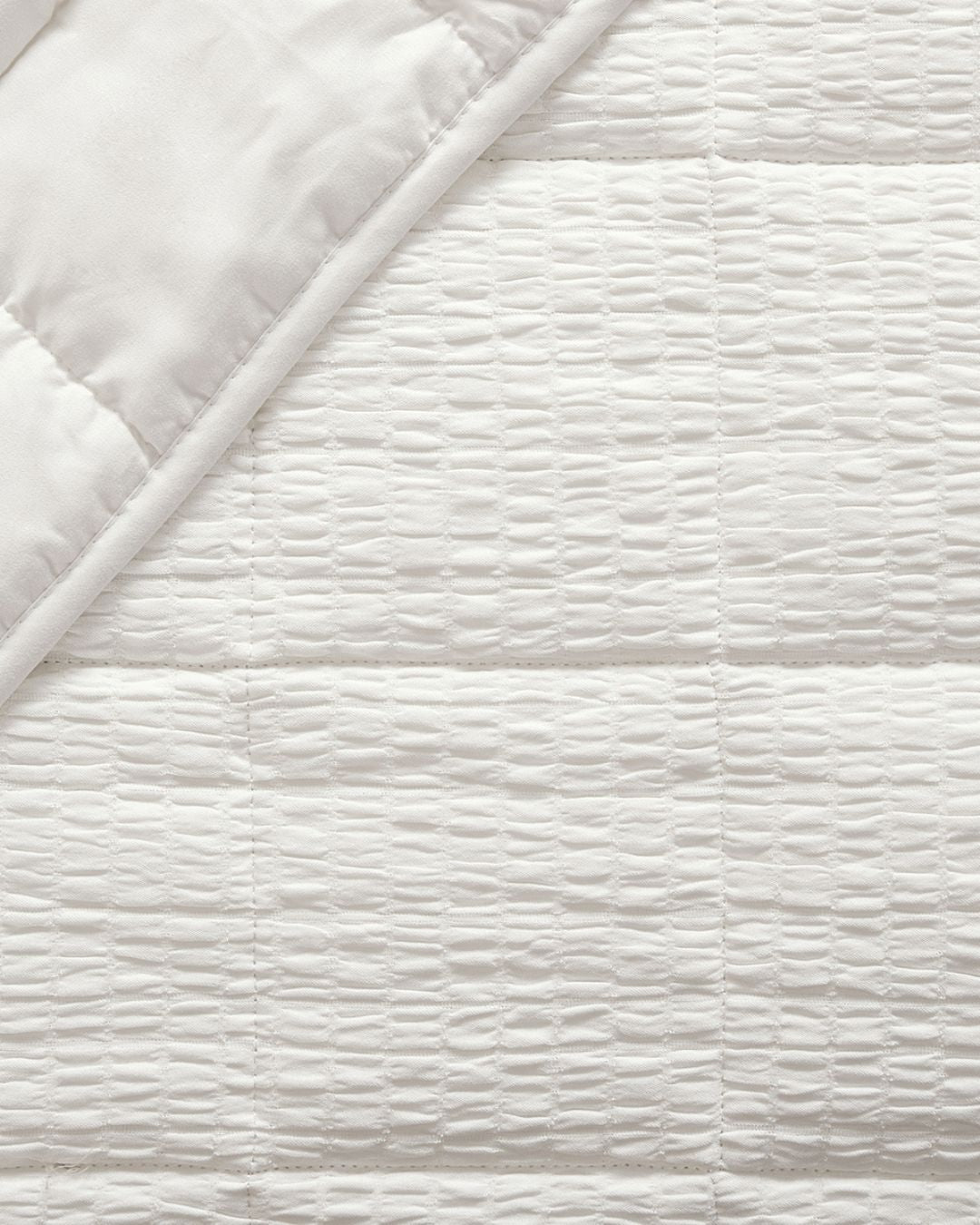 Close-up of a white quilted bedding fabric with a textured, wavy stitched pattern and a folded corner showing the smooth underside.