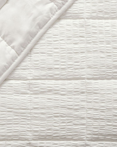 Close-up of a white quilted bedding fabric with a textured, wavy stitched pattern and a folded corner showing the smooth underside.