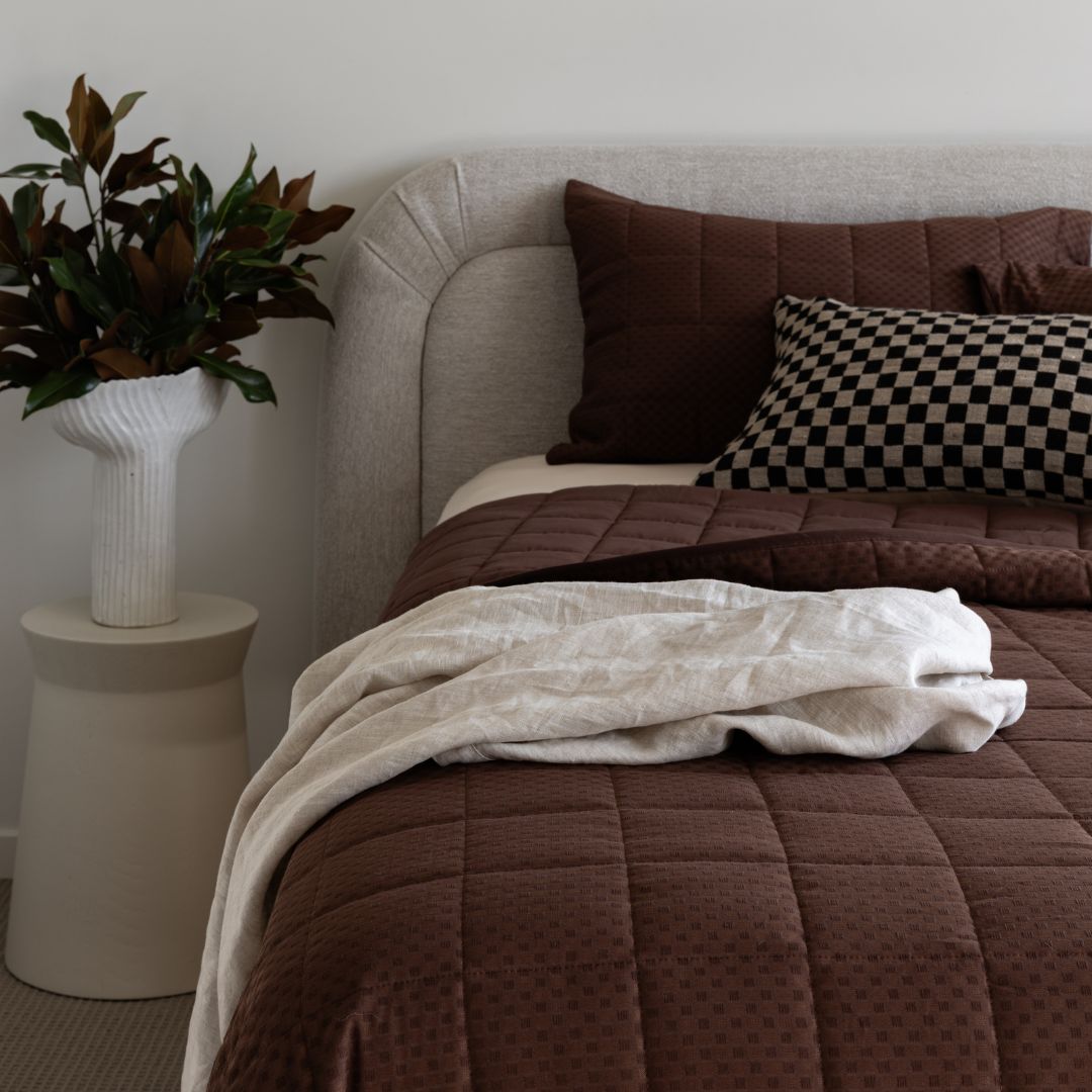 Bed with brown bedding and pillows, next to a plant on a side table.