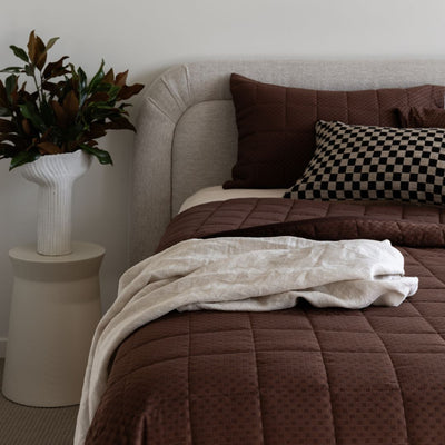 Bed with brown bedding and pillows, next to a plant on a side table.