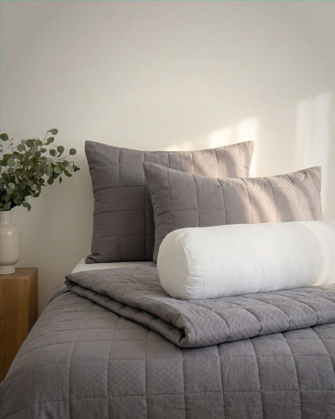 Gray quilted comforter and pillows on a bed with a white wall and plant in the background