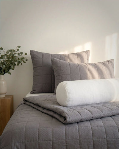Gray quilted comforter and pillows on a bed with a white wall and plant in the background